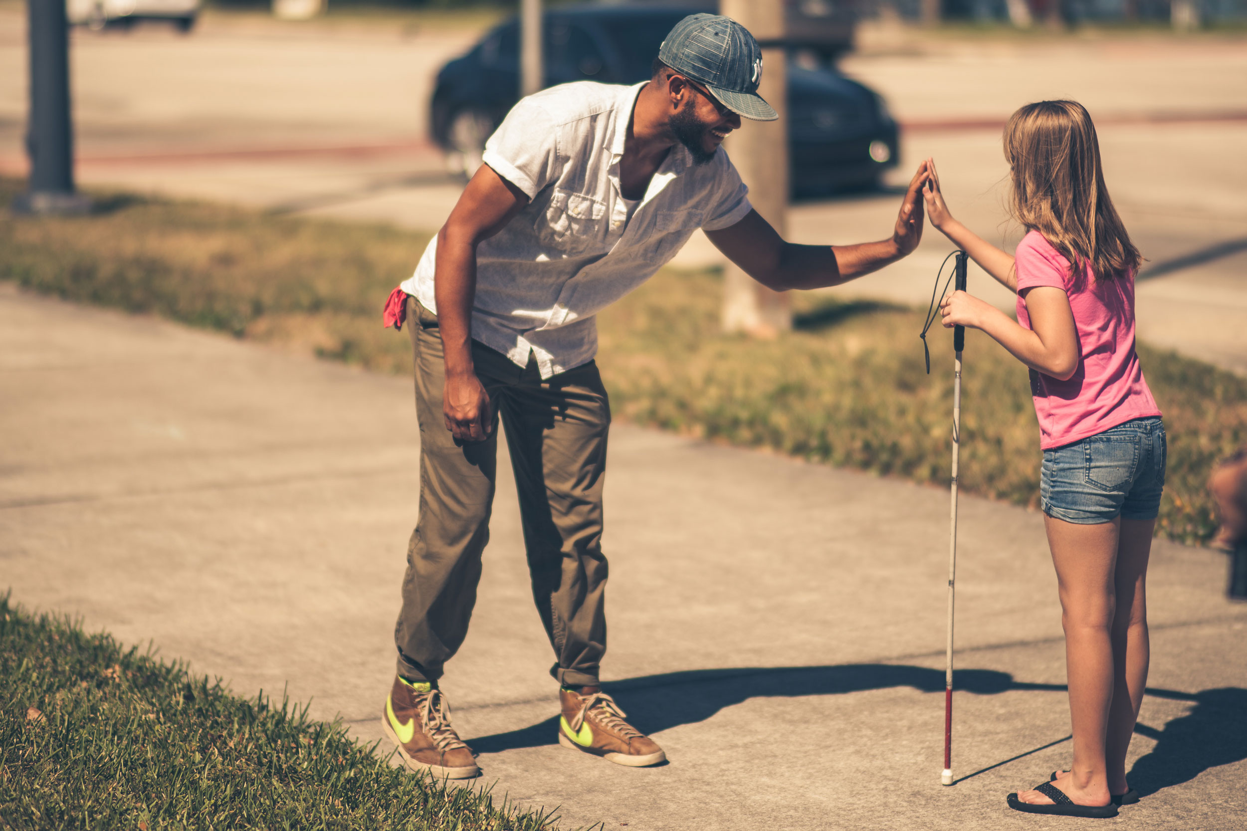 a child who is blind with a white cane in one hand and giving a high five with her other hadn to a person she is passing as she walks on a sidewalk